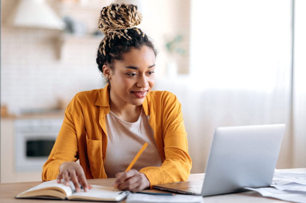 Focused stylish female student studying remotely from home, using a laptop, taking notes on notepad during online lesson, e-learning concept, smiling