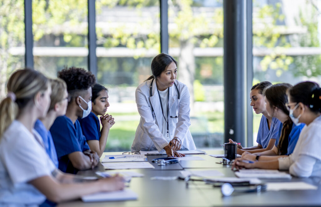 A small group of medical student residents gather around a boardroom table to to meet with their medical team lead. They each have cases out on front of them as they work together collaboratively to discuss each one. They are each dressed professionally in medical scrubs and are listening attentively to the doctor leading the meeting at the head of the table.